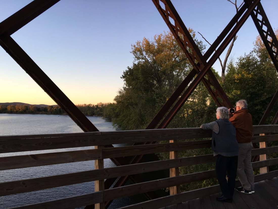 An older couple gazes out over a river from a railroad bridge, under a clear blue evening sky.